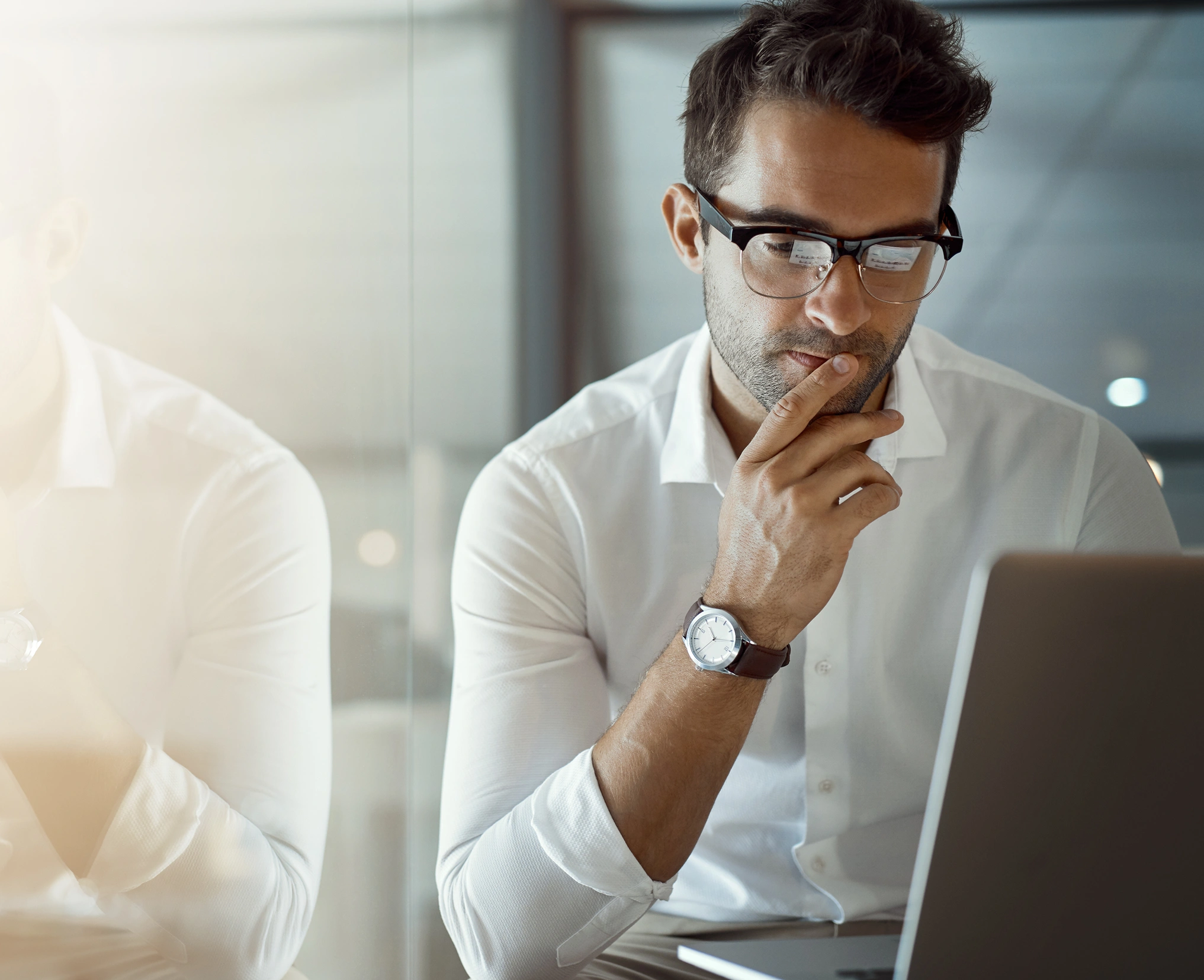 Man in white business shirt and glasses looking intently down at his laptop next to glass window