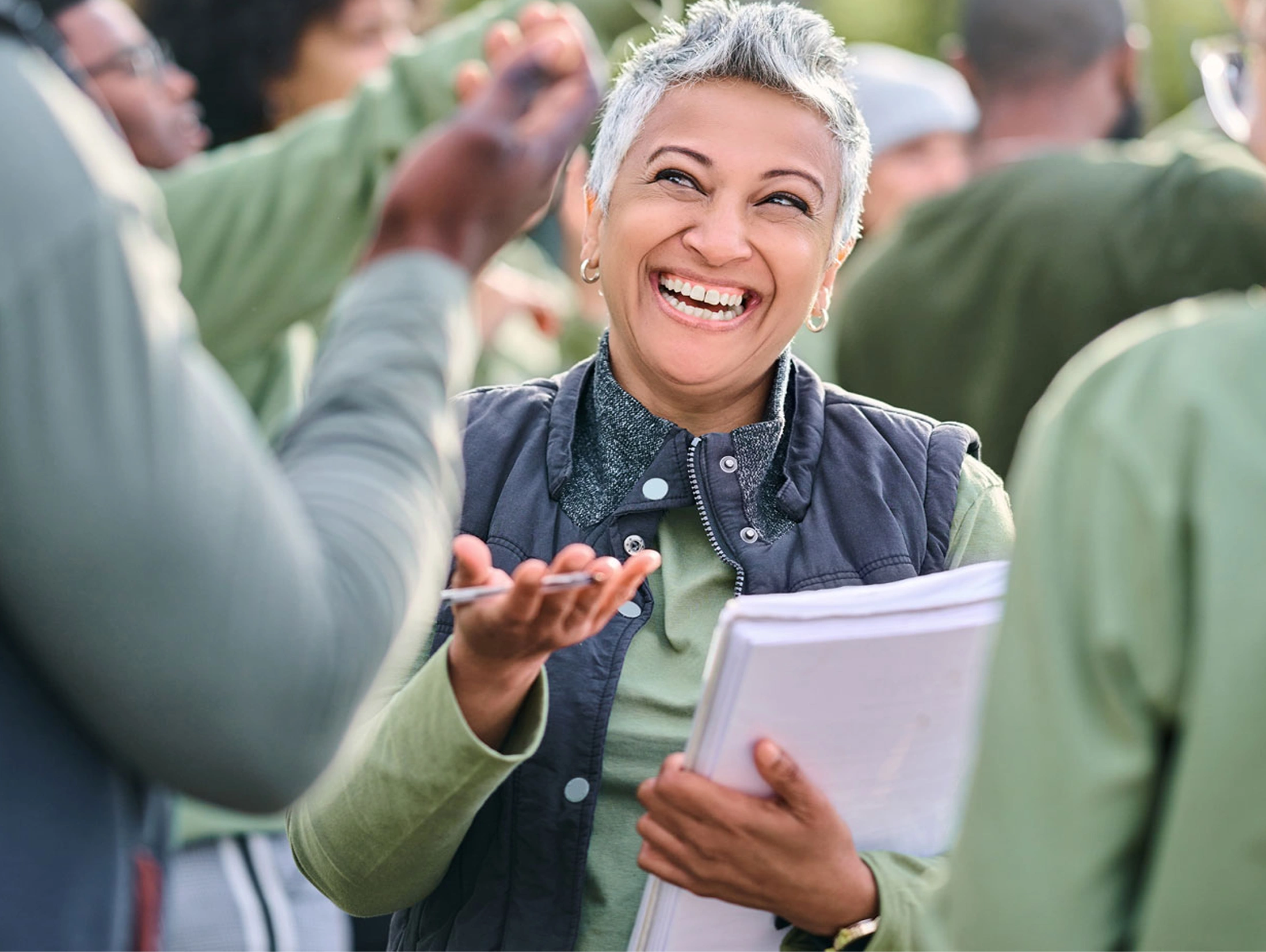 Woman in green shirt and vest, holding papers and clipboard, laughing and talking to other volunteers