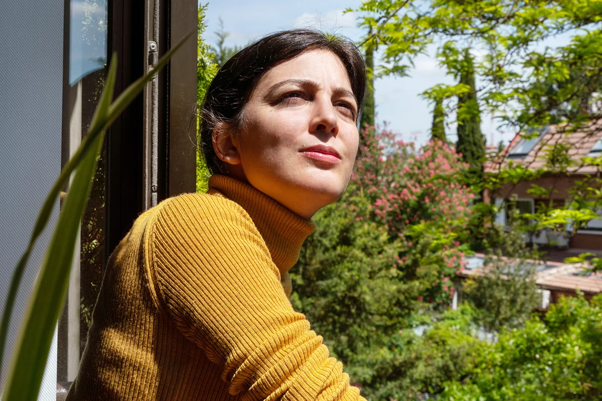 Person standing near a window, looking out at greenery in a calm, reflective outdoor setting.