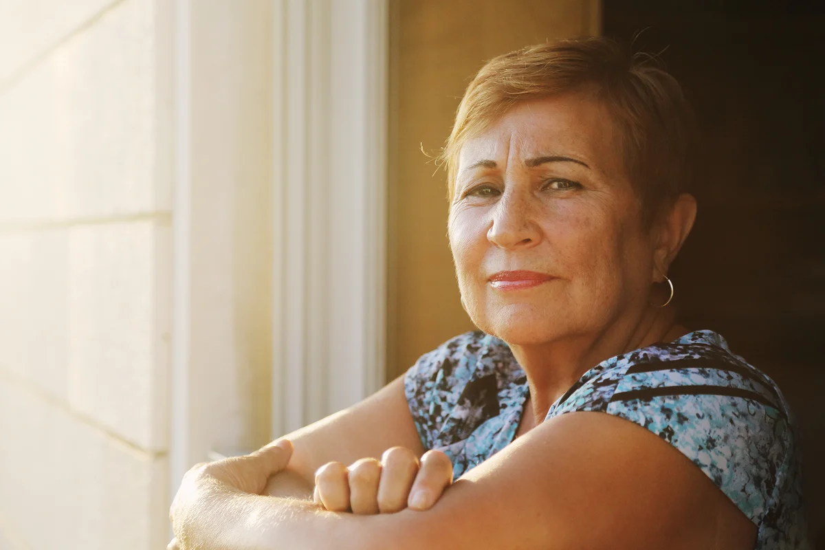 Woman seated by a window, looking thoughtfully toward the camera in a quiet, reflective moment.