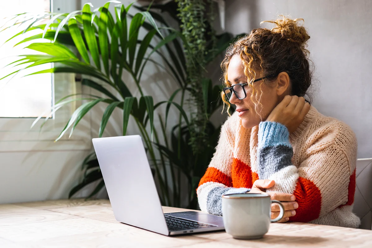 Woman in cozy sweater looking at laptop with coffee mug next to her